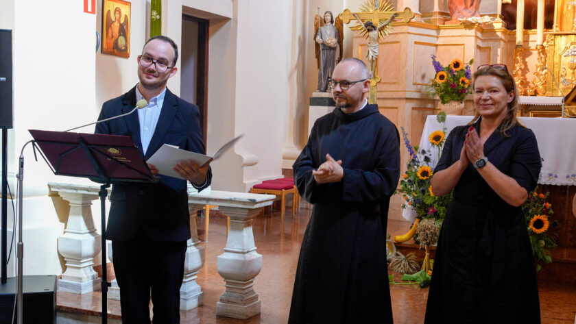 Mężczyzna w ciemnej marynarce stoi przy mikrofonie z otwartą teczką w dłoniach, a obok niego brat zakonny w czarnym habicie i kobieta w ciemnej sukience biją brawo. Scena odbywa się przed ołtarzem podczas jubileuszu warszawskich dzieł bonifraterskich. W tle widoczne są krucyfiks oraz dekoracje kwiatowe ze słonecznikami.