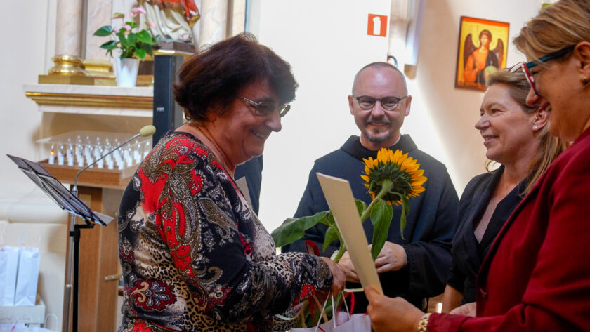 Uśmiechnięta kobieta otrzymuje słonecznik i pamiątkowe podziękowanie podczas uroczystości jubileuszowej w kościele. Wręczeniu towarzyszą brat zakonny w czarnym habicie oraz dwie kobiety. W tle widoczny jest fragment wnętrza świątyni, pulpit z mikrofonem oraz świece wotywne.