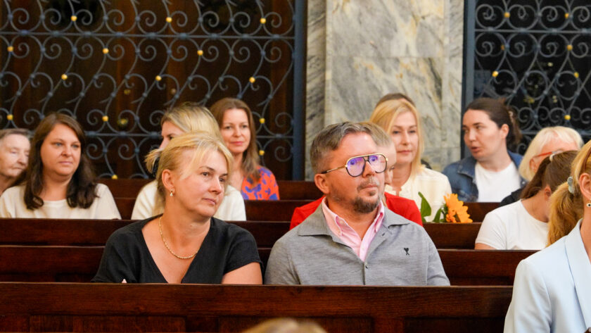 Uczestnicy jubileuszu warszawskich dzieł bonifraterskich siedzą w drewnianych ławkach kościoła i obserwują przebieg uroczystości. Na pierwszym planie widoczni są kobieta w czarnym stroju oraz mężczyzna w okularach, różowej koszuli i szarym swetrze. Za nimi siedzą kolejni uczestnicy spotkania, a w tle widoczne są ozdobne metalowe kraty i marmurowe elementy wnętrza świątyni.