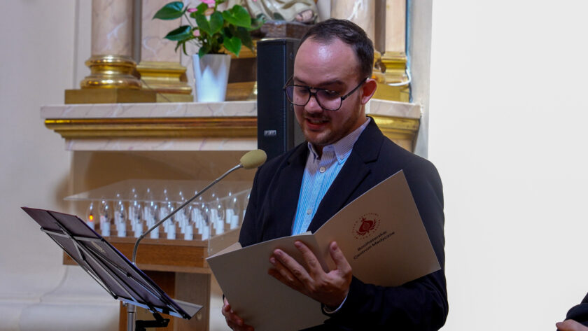 Mężczyzna w okularach i ciemnej marynarce przemawia do mikrofonu podczas uroczystości jubileuszowej w kościele. W dłoniach trzyma otwartą teczkę z logo Bonifraterskiego Centrum Medycznego. Obok znajduje się pulpit na nuty, a w tle fragment ołtarza, kwiaty i świece wotywne.