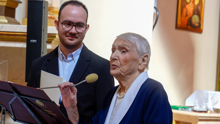 Starsza kobieta przemawiająca do mikrofonu podczas jubileuszowej uroczystości. Ubrana jest w granatowy żakiet z jasnym kołnierzem i perłowy naszyjnik. Za nią stoi młody mężczyzna w ciemnej marynarce i okularach, trzymający dokumenty. W tle widoczne jest wnętrze kościoła oraz elementy wyposażenia ołtarza.