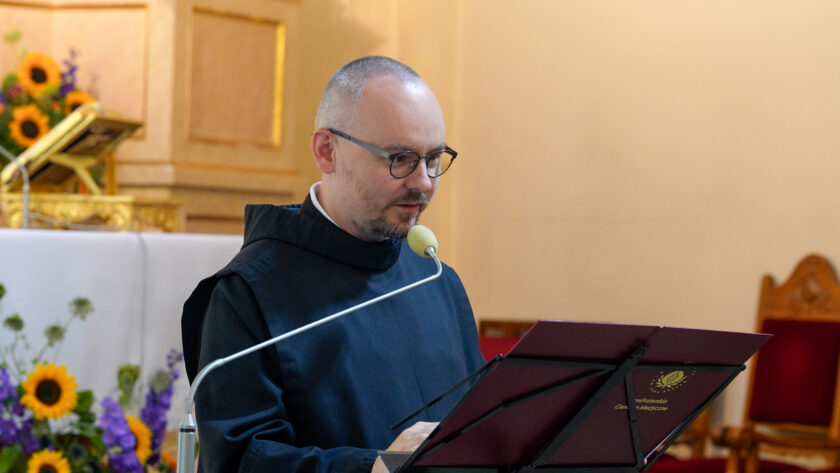 Brat bonifrater w czarnym habicie i okularach przemawiający przy mikrofonie, stojąc za pulpitem z logo Bonifraterskiego Centrum Medycznego. Mężczyzna spogląda na znajdujące się przed nim materiały. W tle widoczny jest fragment ołtarza, kompozycja kwiatowa ze słonecznikami oraz drewniane krzesło.