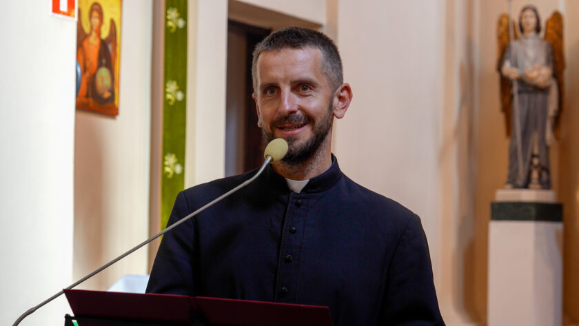 Uśmiechnięty duchowny w czarnym stroju z koloratką przemawiający do mikrofonu podczas uroczystości w kościele. Przed nim znajduje się pulpit, a w tle widoczne są jasne kolumny, zielona dekoracyjna tkanina, obraz religijny oraz figura anioła.