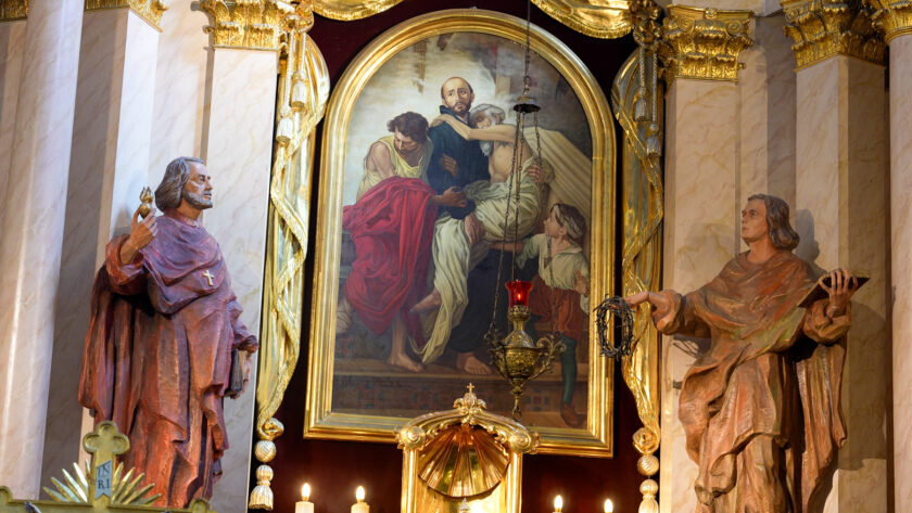 Bogato zdobiony fragment wnętrza kościoła Bonifratrów. W centrum znajduje się duży obraz religijny w złotej, łukowatej ramie, przedstawiający scenę pomocy osobie chorej lub potrzebującej. Po obu stronach obrazu stoją drewniane figury świętych, a przed nim zawieszona jest ozdobna czerwona lampka. Całość otaczają złocone elementy ołtarza i jasne marmoryzowane kolumny.