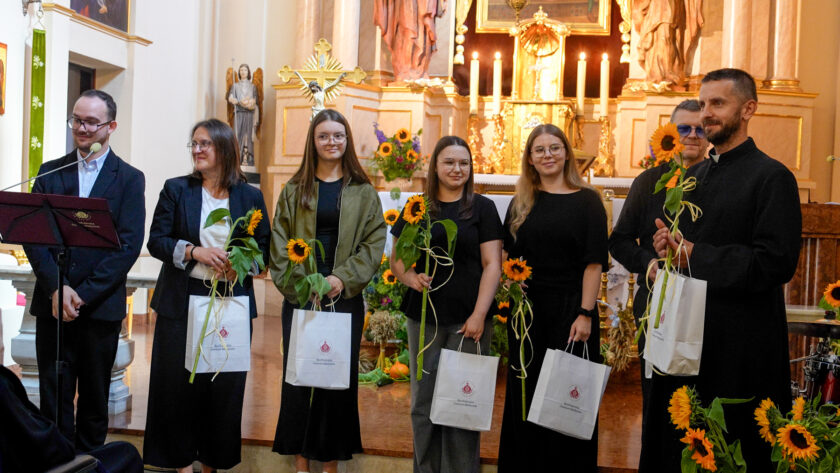 Grupa uczestników uroczystości stojąca przed ołtarzem kościoła. W centrum znajdują się cztery kobiety, które wcześniej występowały wokalnie; trzymają pojedyncze słoneczniki oraz białe torby z logo Bonifraterskiego Centrum Medycznego. Po lewej stronie stoi prowadzący uroczystość przy pulpicie, a po prawej dwóch mężczyzn, w tym duchowny trzymający słonecznik i torbę z upominkiem. W tle widoczny jest bogato zdobiony ołtarz i dekoracje ze słoneczników.