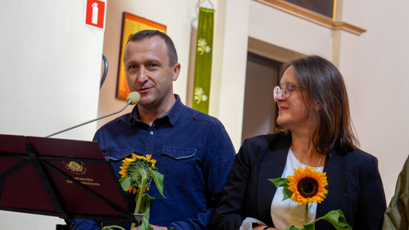 Mężczyzna w granatowej koszuli przemawiający do mikrofonu, stojąc obok kobiety w ciemnej marynarce i okularach. Oboje trzymają duże słoneczniki otrzymane podczas uroczystości. Przed nimi znajduje się pulpit z logo Bonifraterskiego Centrum Medycznego, a w tle widoczne są elementy wystroju kościoła.