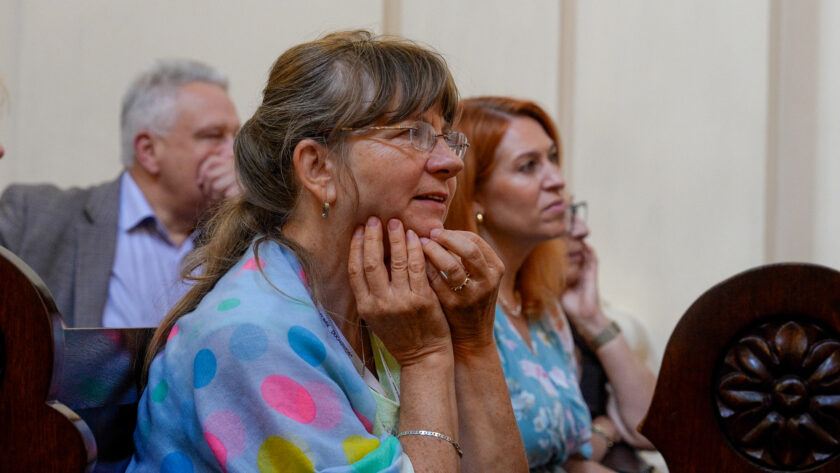 Kobieta w okularach siedząca w kościelnej ławie i z uwagą obserwująca przebieg uroczystości. Ma związane włosy i kolorową chustę w duże niebieskie, różowe i zielone koła, a dłonie opiera na brodzie. Za nią siedzą kolejni uczestnicy wydarzenia, w tym mężczyzna i kobieta, również zwróceni w stronę miejsca, w którym odbywa się uroczystość.
