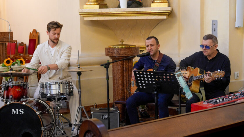 Trzech muzyków występujących podczas jubileuszowego spotkania w kościele. Po lewej mężczyzna gra na perkusji, pośrodku muzyk śpiewa do mikrofonu i gra na gitarze basowej, a po prawej mężczyzna gra na gitarze akustycznej. Przed muzykami znajdują się statywy, instrumenty i sprzęt nagłośnieniowy.