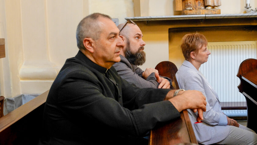 Uczestnicy jubileuszowego spotkania siedzący w kościelnych ławkach i obserwujący wydarzenie. Na pierwszym planie mężczyzna w czarnym stroju, za nim brodaty mężczyzna w okularach, a po prawej kobieta w jasnym stroju. Wnętrze utrzymane jest w jasnej, ciepłej kolorystyce.