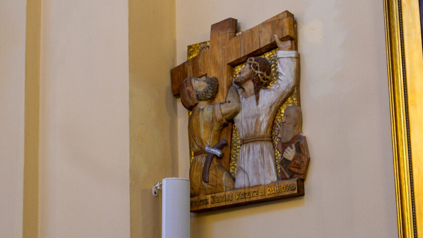 Drewniana płaskorzeźba przedstawiająca jedną ze stacji Drogi Krzyżowej, zawieszona na jasnej ścianie kościoła. Scena przedstawia Jezusa z krzyżem w otoczeniu dwóch postaci. Pod płaskorzeźbą widoczny jest fragment głośnika.
