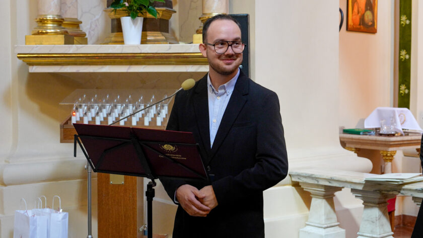 Mężczyzna w okularach i ciemnym garniturze stoi przy pulpicie z mikrofonem we wnętrzu kościoła. Uśmiecha się i spogląda w bok. W tle widoczne są elementy ołtarza, świece oraz jasne, złocone detale wyposażenia świątyni.