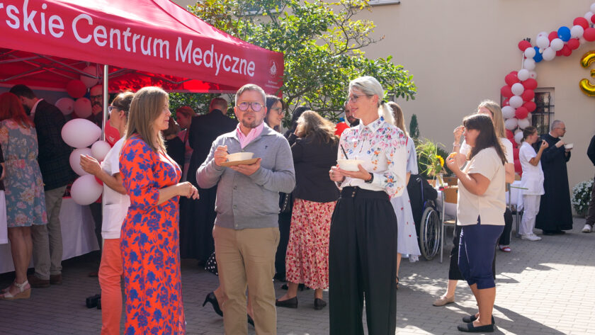 Uczestnicy jubileuszu rozmawiają i częstują się jedzeniem podczas spotkania na dziedzińcu. Na pierwszym planie stoją dwie kobiety i mężczyzna trzymający miskę. W tle znajduje się czerwony namiot Bonifraterskiego Centrum Medycznego, biało-czerwone dekoracje z balonów oraz pozostali uczestnicy wydarzenia.