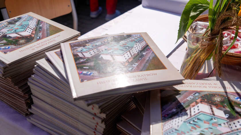Stosy jubileuszowych publikacji zatytułowanych „Historia Bonifratrów w Warszawie” ułożone na stole. Na okładce znajduje się fotografia kompleksu budynków z lotu ptaka. Obok widoczna jest roślinna dekoracja w szklanym naczyniu oraz fragment koszyka z drobnymi upominkami.