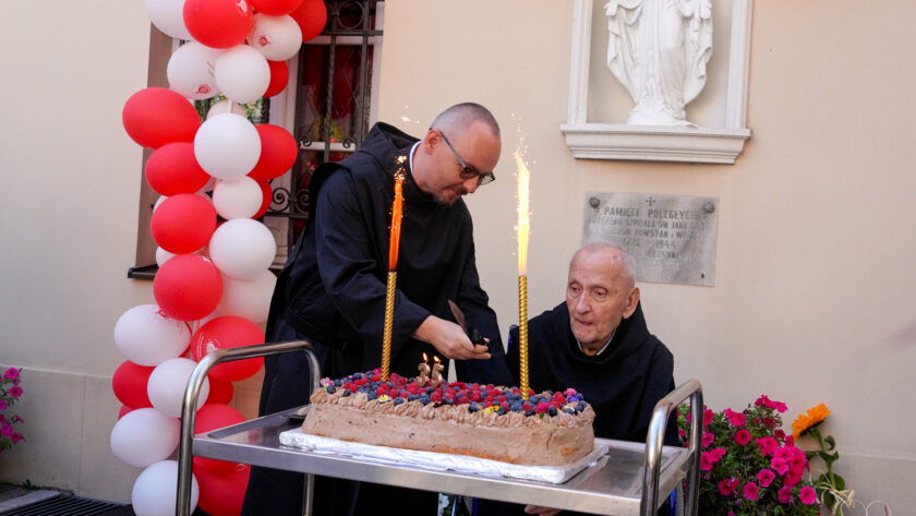 Na dziedzińcu przed budynkiem dwóch bonifratrów stoi przy dużym jubileuszowym torcie udekorowanym owocami, świeczkami w kształcie liczby „35” oraz dwiema wysokimi racami tortowymi. Jeden z zakonników pochyla się nad tortem, drugi obserwuje przygotowania. W tle widoczna jest biało-czerwona dekoracja z balonów oraz fragment elewacji budynku.