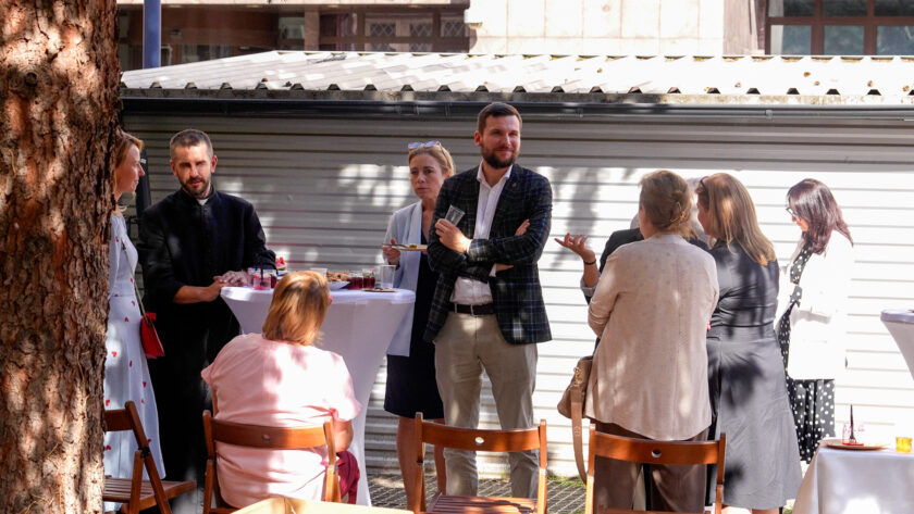Uczestnicy jubileuszowego spotkania rozmawiają na dziedzińcu przy wysokich stolikach. W centrum kadru stoją mężczyzna w marynarce oraz kobieta trzymająca talerzyk, po lewej widoczny jest duchowny w czarnym stroju. Wokół nich znajdują się pozostali goście wydarzenia, a część osób korzysta z przygotowanego poczęstunku.