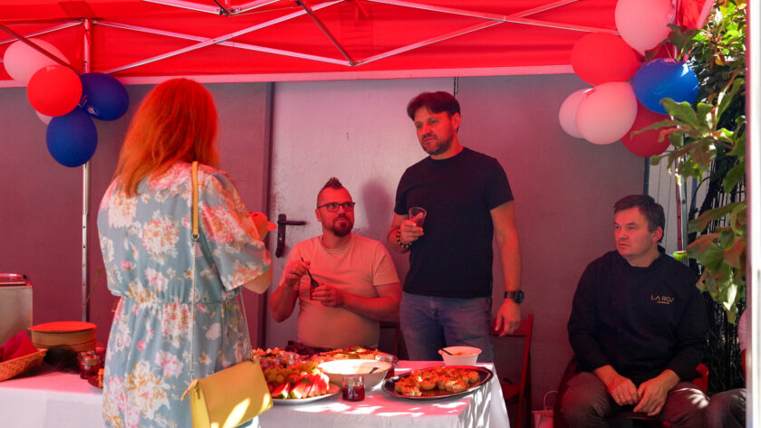 Uczestnicy spotkania korzystają z poczęstunku przygotowanego pod czerwonym namiotem udekorowanym biało-czerwonymi i niebieskimi balonami. Przy stole z przekąskami znajdują się trzy osoby, a na pierwszym planie stoi kobieta w jasnej sukience w kwiaty.