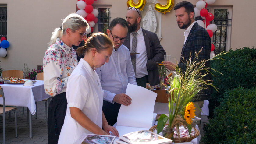 Grupa uczestników jubileuszu zgromadzona przy stole z księgą pamiątkową i publikacjami „Historia Bonifratrów w Warszawie”. Na pierwszym planie kobieta w białym stroju porządkuje materiały, a obok niej trzy osoby oglądają otwartą księgę. Stół udekorowany jest kompozycją ze słonecznikiem i ozdobnymi trawami.