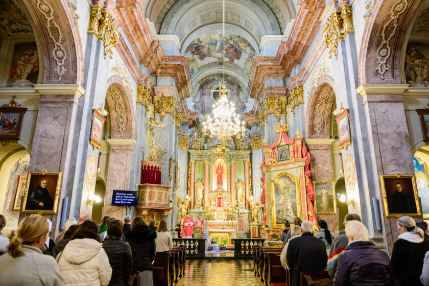 Widok na wnętrze zabytkowego kościoła podczas Mszy Świętej z udziałem bonifratrów, duchowieństwa i zgromadzonych wiernych. Uczestnicy stoją w drewnianych ławkach, zwróceni w kierunku ołtarza, przy którym znajdują się kapłani w czerwonych szatach liturgicznych. Wnętrze świątyni zdobią marmoryzowane kolumny, złocone ornamenty, obrazy religijne, freski na sklepieniu oraz okazały kryształowy żyrandol.