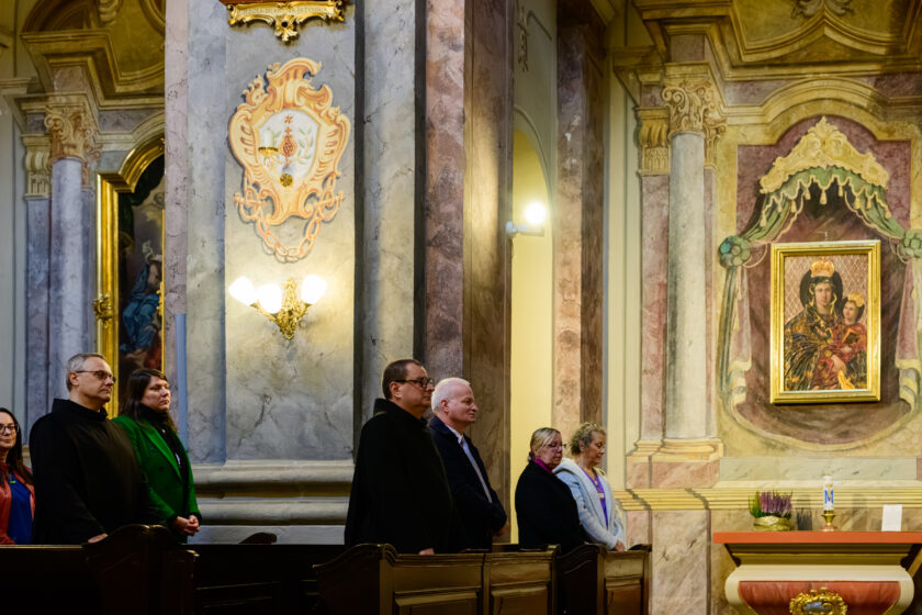 Uczestnicy uroczystości religijnej stoją w ławkach zabytkowego kościoła. Na pierwszym planie widoczni są dwaj bonifratrzy w czarnych habitach, mężczyzna w ciemnym garniturze oraz kilka kobiet. Po prawej stronie znajduje się boczny ołtarz z obrazem Matki Bożej z Dzieciątkiem Jezus w złoconej ramie. Ściany świątyni zdobią marmoryzowane kolumny, dekoracyjne ornamenty i ozdobne kinkiety.