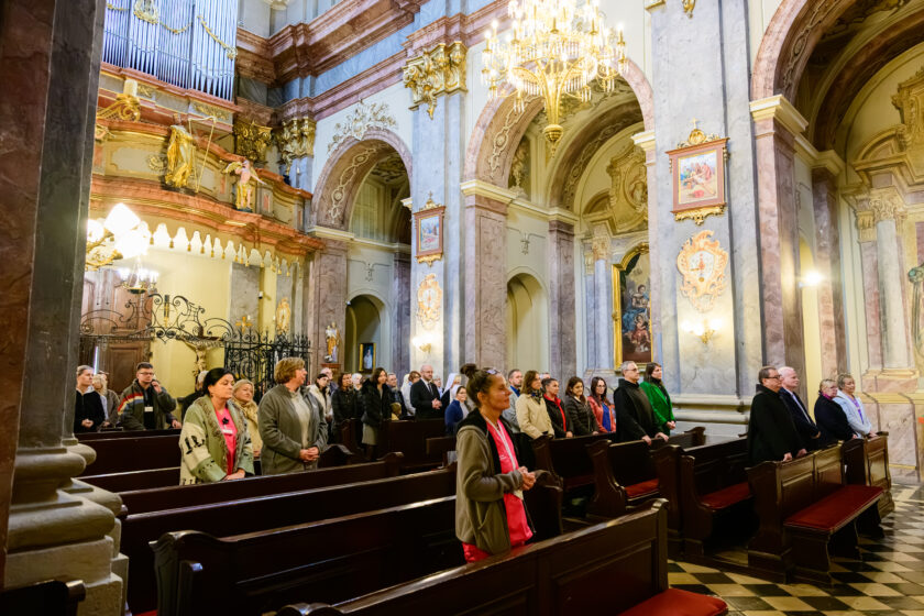 Boczny widok na zgromadzonych wiernych uczestniczących w uroczystej Mszy Świętej w zabytkowym kościele. Kobiety i mężczyźni w różnym wieku stoją w drewnianych ławkach, niektórzy ze złożonymi dłońmi. Wśród uczestników znajdują się bonifratrzy w czarnych habitach. W tle widoczne są organy piszczałkowe, bogato zdobione kolumny, łuki architektoniczne, obrazy religijne oraz rozświetlony żyrandol.