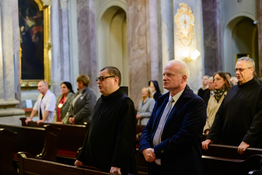 Zbliżenie na uczestników Mszy Świętej stojących w drewnianych ławkach kościoła. Na pierwszym planie widoczny jest bonifrater w czarnym habicie i okularach, obok niego stoi siwowłosy mężczyzna w granatowym garniturze i jasnym krawacie. Po prawej stronie znajduje się kolejny bonifrater w czarnym habicie. W tle widoczni są pozostali uczestnicy uroczystości oraz marmoryzowane kolumny i ozdobne elementy wnętrza świątyni.