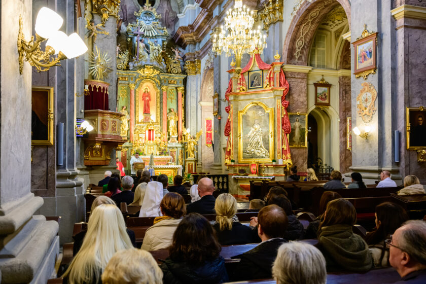 Widok z tylnej części zabytkowego kościoła na uczestników Mszy Świętej siedzących w drewnianych ławkach. Przy głównym ołtarzu znajduje się kapłan w jasnych szatach liturgicznych, a obok niego ministrant w czerwonym stroju. W centrum prezbiterium stoi figura Matki Bożej Fatimskiej. Wnętrze świątyni zdobią złocone ołtarze, obrazy religijne, marmoryzowane kolumny oraz duży kryształowy żyrandol.
