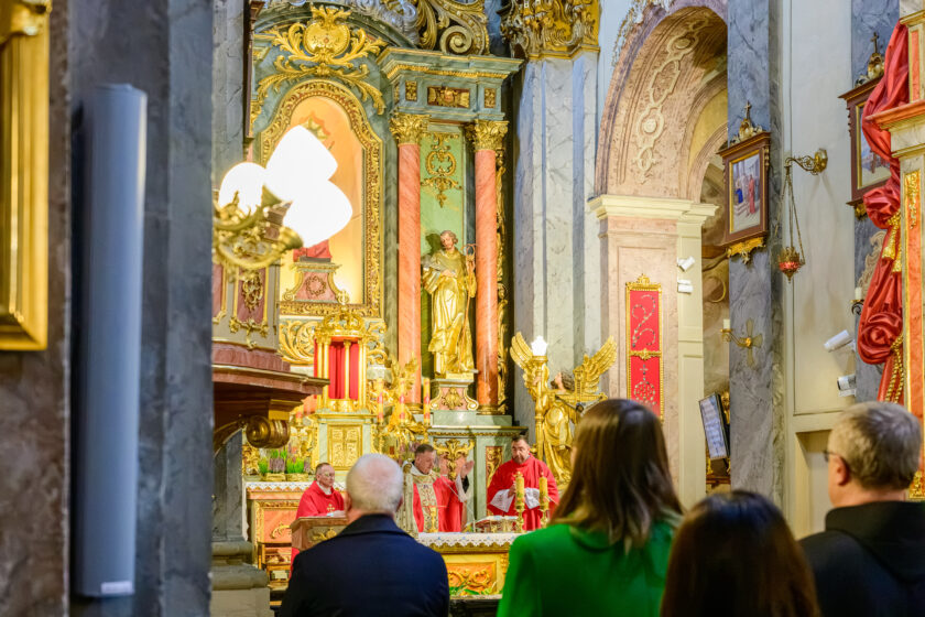 Widok z bocznej części na prezbiterium zabytkowego kościoła podczas sprawowania Mszy Świętej. Przy ołtarzu stoją trzej kapłani w czerwonych szatach liturgicznych. Jeden z nich unosi prawą dłoń w geście modlitwy. Na pierwszym planie, od tyłu, widoczni są uczestnicy uroczystości, w tym kobieta w zielonym płaszczu i mężczyźni w ciemnych ubraniach. W tle znajduje się bogato zdobiony złoceniami ołtarz oraz figury aniołów.