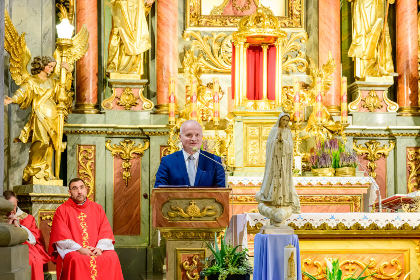 Siwowłosy mężczyzna ubrany w granatowy garnitur, białą koszulę i jasny krawat przemawia przy drewnianej ambonie w zabytkowym kościele. Po lewej stronie siedzi kapłan w czerwonym ornacie ze złotymi zdobieniami. Za przemawiającym znajduje się bogato dekorowany ołtarz z figurami aniołów, złoconym tabernakulum, świecami i kompozycjami z wrzosów. Na pierwszym planie po prawej stronie stoi figura Matki Bożej Fatimskiej z różańcem.