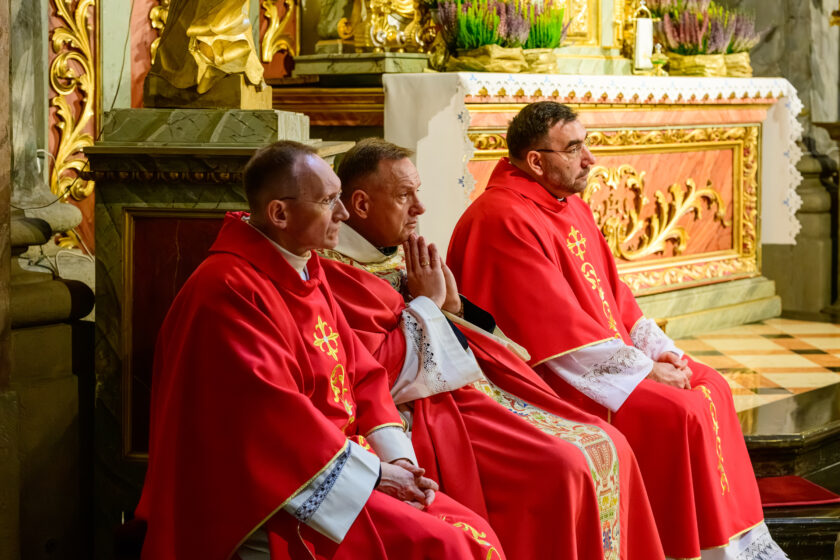 Trzech kapłanów uczestniczy w uroczystej Mszy Świętej w zabytkowym kościele. Duchowni siedzą obok siebie w prezbiterium, ubrani w czerwone szaty liturgiczne. Kapłan pośrodku, w jasnej, bogato haftowanej szacie, trzyma dłonie złożone do modlitwy przy twarzy. Dwaj pozostali duchowni siedzą po jego bokach, zwróceni w stronę ołtarza. W tle widoczne są złocone ornamenty, fragment ołtarza oraz dekoracje z wrzosów.