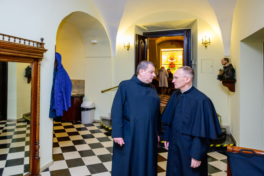 Dwaj duchowni w czarnych strojach zakonnych rozmawiają w jasnym korytarzu zabytkowego budynku. Mężczyzna po lewej stronie, ubrany w czarny habit, zwraca się do duchownego w okularach, noszącego czarną sutannę z guzikami i krótką peleryną naramienną. Pomieszczenie ma sklepienie kolebkowe, jasne ściany i czarno-białą posadzkę ułożoną w szachownicę. W tle widoczne są otwarte drewniane drzwi, kolorowy witraż oraz figura zakonnika umieszczona na ściennej półce.