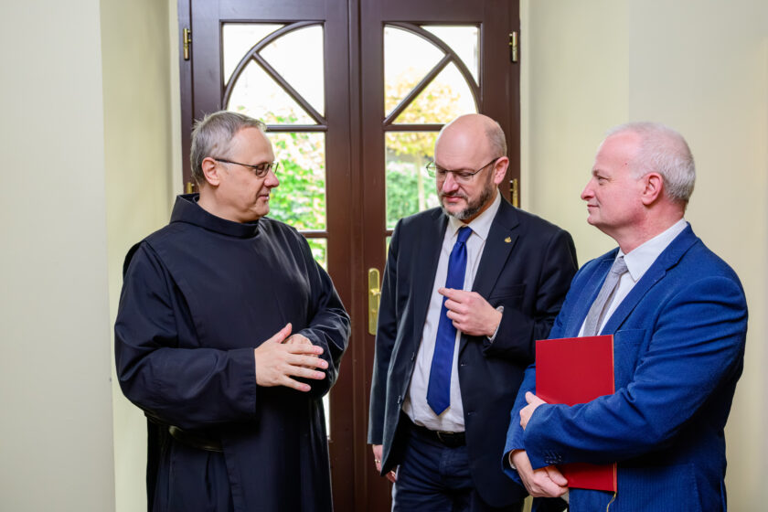Trzech mężczyzn prowadzi rozmowę w jasnym pomieszczeniu przed przeszklonymi drewnianymi drzwiami. Po lewej stronie stoi siwowłosy bonifrater w czarnym habicie i okularach, gestykulujący podczas rozmowy. Pośrodku znajduje się łysiejący mężczyzna z brodą i okularami, ubrany w ciemny garnitur, białą koszulę i niebieski krawat. Po prawej stronie stoi siwowłosy mężczyzna w niebieskiej marynarce i jasnym krawacie, trzymający pod pachą czerwoną teczkę. Za przeszklonymi drzwiami widoczna jest zieleń.
