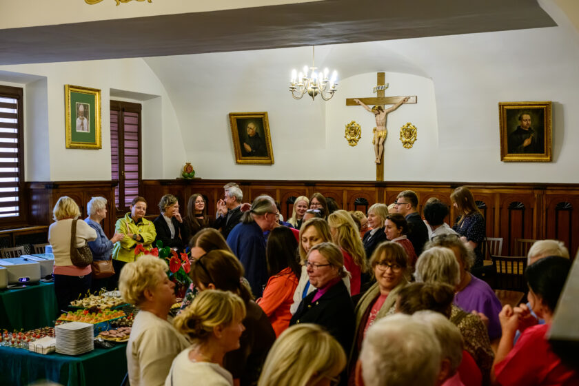 Uczestnicy spotkania zgromadzeni w zabytkowej sali z drewnianą boazerią i sklepionym sufitem. Kobiety i mężczyźni w różnym wieku stoją w kilku grupach, rozmawiając ze sobą. Na pierwszym planie po lewej stronie znajduje się stół nakryty zielonym obrusem, na którym przygotowano różnorodne przekąski, małe porcje deserów i talerze. Na tylnej ścianie sali umieszczony jest duży krucyfiks z figurą ukrzyżowanego Chrystusa, a po jego bokach wiszą obrazy religijne w złoconych ramach. Pomieszczenie oświetla ozdobny żyrandol.