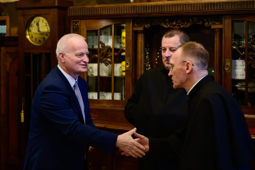 Siwowłosy mężczyzna w granatowym garniturze, białej koszuli i jasnym krawacie wita się z duchownym w czarnej sutannie i okularach. Mężczyźni podają sobie prawe dłonie podczas spotkania w pomieszczeniu z zabytkowymi drewnianymi meblami. Między nimi, nieco w głębi, stoi bonifrater w czarnym habicie, obserwujący powitanie. W tle znajdują się przeszklone kredensy z naczyniami oraz duży drewniany zegar z okrągłą tarczą.