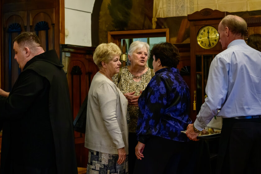 Grupa uczestników uroczystości rozmawia w pomieszczeniu wyposażonym w zabytkowe drewniane meble. W centrum zdjęcia stoją trzy kobiety: starsza kobieta w jasnym swetrze i wzorzystej spódnicy, siwowłosa kobieta w beżowo-czarnej bluzce oraz kobieta w granatowej bluzce z niebieskim kwiatowym wzorem, odwrócona plecami do aparatu. Po lewej stronie znajduje się bonifrater w czarnym habicie, a po prawej mężczyzna w jasnoniebieskiej koszuli, trzymający tacę z poczęstunkiem. W tle widoczny jest drewniany zegar stojący oraz przeszklone kredensy.