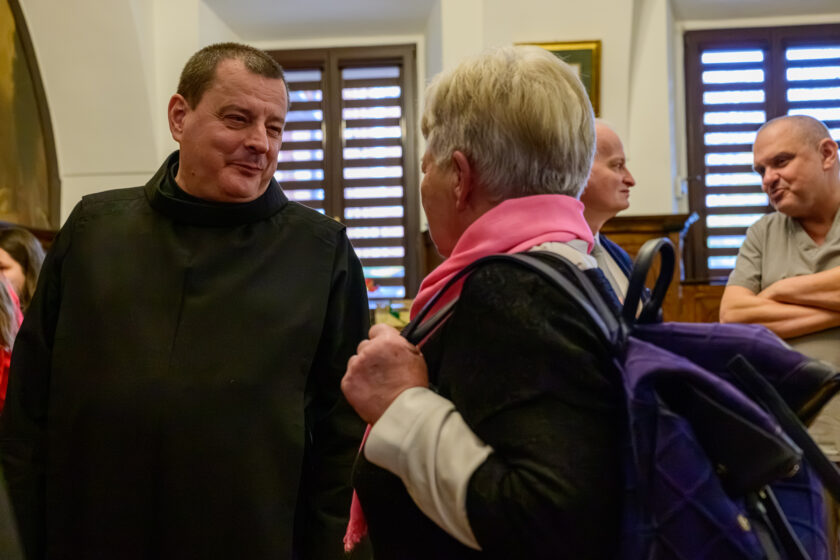 Bonifrater w czarnym habicie rozmawia z siwowłosą kobietą podczas spotkania w zabytkowej sali. Zakonnik stoi po lewej stronie kadru, uśmiechając się do rozmówczyni. Kobieta, odwrócona częściowo plecami do aparatu, ubrana jest w ciemny strój z jasnymi mankietami i różowym szalem. Na ramionach ma granatowy plecak, którego pasek przytrzymuje dłonią. W tle widoczni są dwaj mężczyźni, w tym jeden w jasnoszarym stroju medycznym, opierający skrzyżowane ręce na blacie. Pomieszczenie zdobi drewniana boazeria, a światło wpada przez okna z poziomymi roletami.