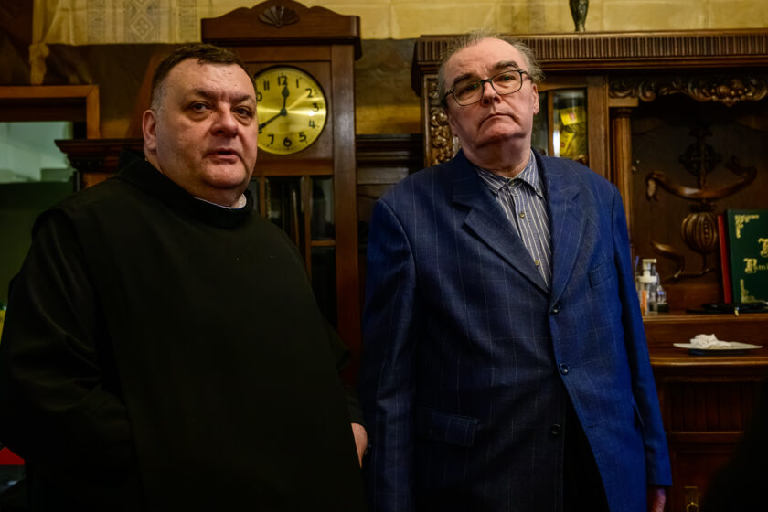 Dwaj mężczyźni pozują do zdjęcia w zabytkowym pomieszczeniu wyposażonym w drewniane meble. Po lewej stronie stoi bonifrater w czarnym habicie, z krótkimi ciemnymi włosami. Obok niego znajduje się starszy mężczyzna z okularami, ubrany w niebieską marynarkę w delikatną kratę oraz koszulę w pionowe paski. Obaj patrzą w kierunku aparatu. W tle widoczny jest wysoki drewniany zegar z okrągłą tarczą oraz bogato rzeźbiony, przeszklony kredens z naczyniami i dekoracjami.