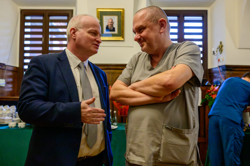 Dwaj mężczyźni rozmawiają podczas spotkania w zabytkowej sali z drewnianą boazerią. Po lewej stronie stoi siwowłosy mężczyzna w granatowym garniturze, białej koszuli i jasnym krawacie, który gestykuluje prawą dłonią podczas rozmowy. Naprzeciwko niego znajduje się uśmiechnięty mężczyzna z krótko ostrzyżonymi włosami, ubrany w jasnoszary strój medyczny, ze skrzyżowanymi rękami. W tle widoczne są okna z brązowymi roletami, obraz w złoconej ramie oraz stół z poczęstunkiem nakryty zielonym obrusem. Po prawej stronie inny uczestnik spotkania w niebieskim stroju medycznym pochyla się nad stołem.