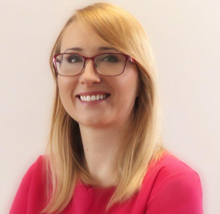 Portret uśmiechniętej kobiety z długimi, prostymi blond włosami oraz czerwono-bordowymi okularami. Kobieta ubrana jest w intensywnie różową bluzkę i patrzy w stronę aparatu. Zdjęcie wykonano od klatki piersiowej w górę na bardzo jasnym, niemal białym tle. Fotografia ma miękkie oświetlenie i lekko rozmyty charakter.