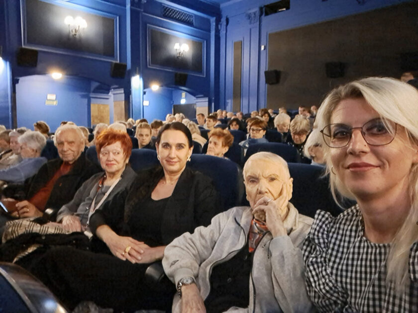 Pięć osób siedzących obok siebie w fotelach na widowni sali kinowej. Od lewej znajdują się starszy mężczyzna, kobieta z krótkimi rudymi włosami, kobieta z ciemnymi włosami, starsza kobieta w jasnej bluzie oraz blondynka w okularach wykonująca selfie. Za nimi widoczne są kolejne rzędy foteli wypełnione widzami. Wnętrze sali ma niebieskie ściany i dekoracyjne oświetlenie.