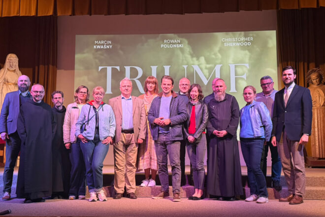 Zdjęcie grupowe przedstawiające trzynaście osób pozujących na scenie przed ekranem, na którym wyświetlany jest plakat filmu „Triumf” z nazwiskami aktorów. Wśród uczestników znajdują się zakonnicy w czarnych habitach, kobiety i mężczyźni w strojach codziennych oraz eleganckich. W centralnej części grupy stoi mężczyzna w ciemnej pikowanej kurtce. Po obu stronach sceny widoczne są rzeźby postaci religijnych, a nad uczestnikami znajduje się brązowo-złota kurtyna.