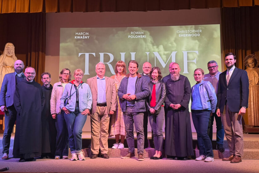 Zdjęcie grupowe przedstawiające trzynaście osób pozujących na scenie przed ekranem, na którym wyświetlany jest plakat filmu „Triumf” z nazwiskami aktorów. Wśród uczestników znajdują się zakonnicy w czarnych habitach, kobiety i mężczyźni w strojach codziennych oraz eleganckich. W centralnej części grupy stoi mężczyzna w ciemnej pikowanej kurtce. Po obu stronach sceny widoczne są rzeźby postaci religijnych, a nad uczestnikami znajduje się brązowo-złota kurtyna.
