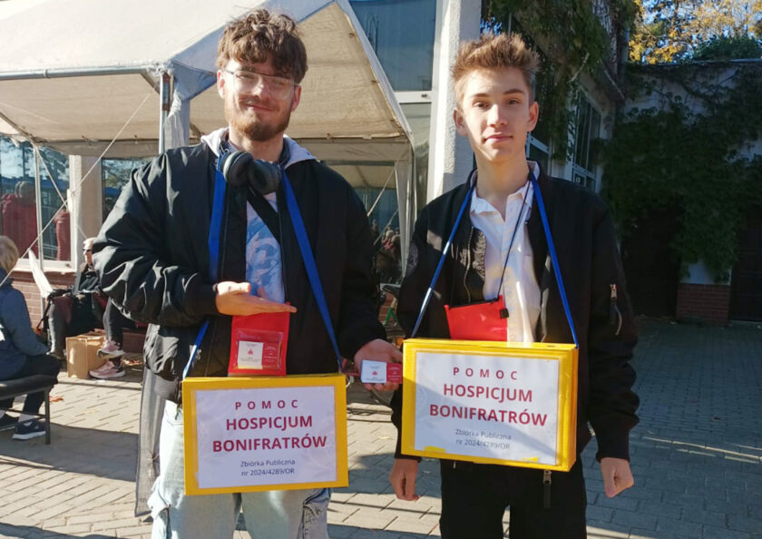 Dwóch młodych wolontariuszy z żółtymi puszkami kwestarskimi na rzecz Hospicjum Bonifratrów. Mężczyźni stoją na alejce w pobliżu cmentarza.