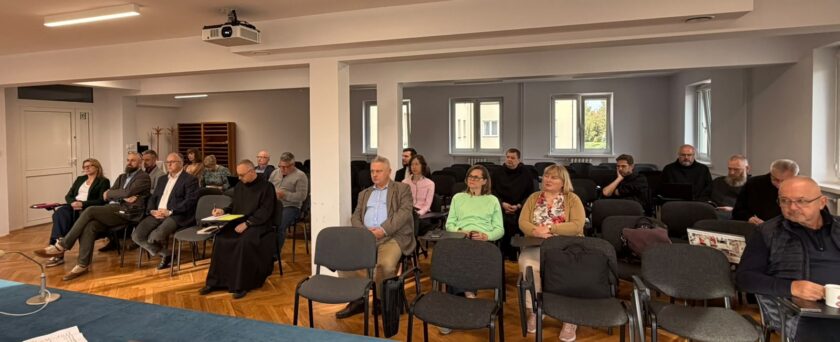 Wśród obecnych znajdują się zakonnicy w czarnych habitach oraz kobiety i mężczyźni w strojach codziennych i formalnych. Część osób trzyma notatniki, dokumenty lub telefony. Na pierwszym planie widoczny jest fragment stołu przykrytego niebieskim obrusem i mikrofon konferencyjny. Sala ma drewnianą podłogę, białe ściany, duże okna oraz projektor zamontowany pod sufitem.