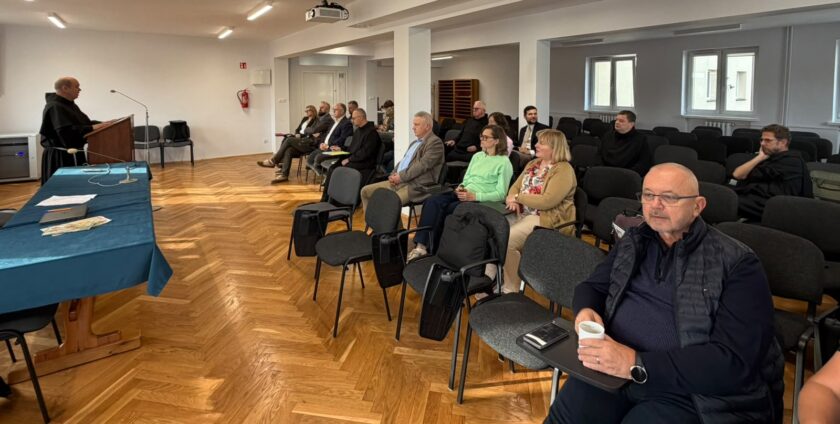 Widok sali konferencyjnej podczas wystąpienia zakonnika w czarnym habicie, stojącego przy drewnianej mównicy z mikrofonem. Przed nim znajduje się stół przykryty niebieskim obrusem, na którym leżą dokumenty i materiały drukowane. Uczestnicy spotkania, w tym zakonnicy oraz osoby świeckie, siedzą na czarnych krzesłach i słuchają przemówienia. Na pierwszym planie po prawej stronie siedzi mężczyzna w okularach i ciemnej kamizelce, trzymający biały kubek. W sali widoczne są drewniana podłoga, białe kolumny, okna oraz projektor sufitowy.
