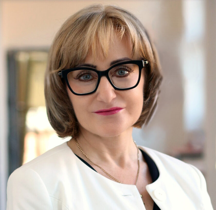 Portret kobiety o krótkich, jasnobrązowych włosach z grzywką, w dużych czarnych okularach. Kobieta patrzy bezpośrednio w obiektyw i lekko się uśmiecha. Ubrana jest w biały żakiet narzucony na czarną bluzkę oraz delikatny złoty łańcuszek. Zdjęcie wykonano od klatki piersiowej w górę. W tle widoczne jest jasne, rozmyte wnętrze.