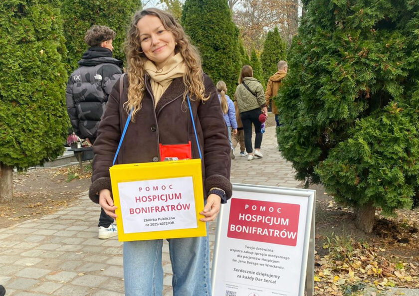 Uśmiechnięta wolontariuszka trzyma żółtą puszkę kwestarską z napisem „Pomoc Hospicjum Bonifratrów”. Obok niej znajduje się tablica informacyjna dotycząca zbiórki.
