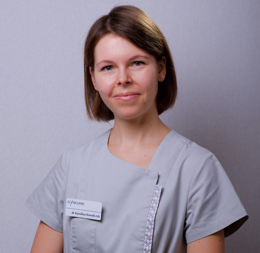 Portret kobiety o krótkich, prostych brązowych włosach, ubranej w jasnoszary strój medyczny. Kobieta stoi na tle jednolitej, jasnoszarej ściany i patrzy w obiektyw z delikatnym uśmiechem. Na lewej stronie stroju ma przypięty biały identyfikator z logo GYNCLINIC oraz napisem „dr Karolina Kowalczyk”. Strój posiada ozdobną, pionową lamówkę z geometrycznym wzorem. Zdjęcie wykonane od pasa w górę.
