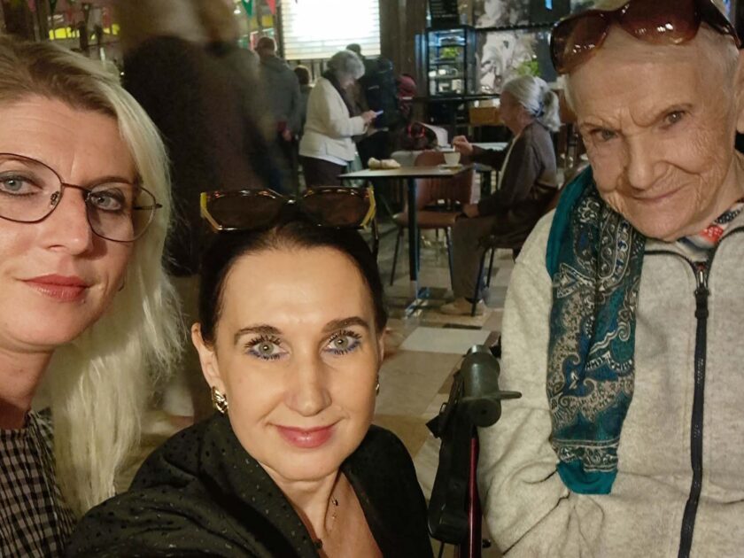 Selfie trzech kobiet wykonane we wnętrzu kawiarni lub restauracji. Po lewej znajduje się blondynka w okularach, pośrodku kobieta z ciemnymi włosami i okularami przeciwsłonecznymi założonymi na głowę, a po prawej starsza kobieta siedząca przy balkoniku, ubrana w jasną bluzę i niebiesko-zielony wzorzysty szalik. W tle przy stolikach siedzą inne osoby.