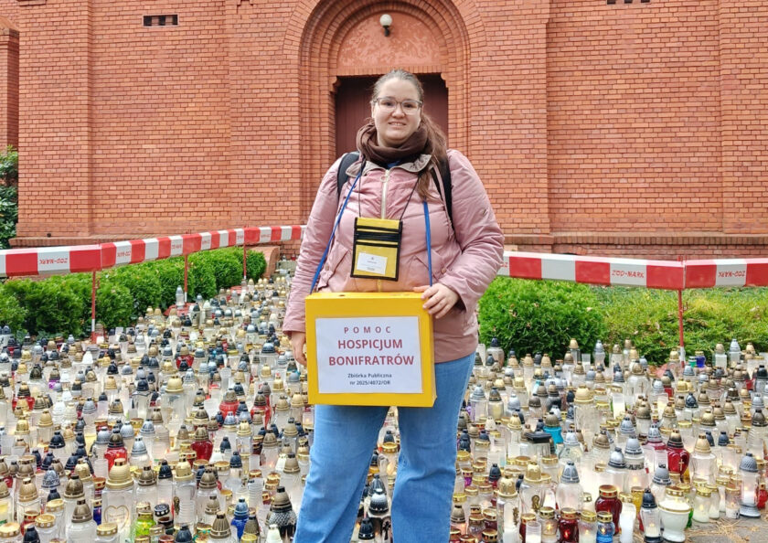 Wolontariuszka z żółtą puszką kwestarską stoi przed ceglaną kaplicą, w otoczeniu licznych zniczy. Bierze udział w zbiórce na rzecz Hospicjum Bonifratrów.