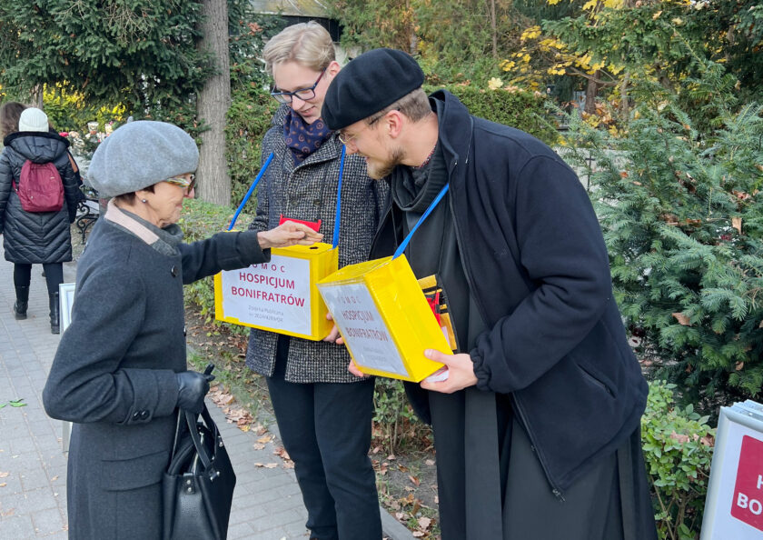 Starsza kobieta przekazuje datek do puszki kwestarskiej trzymanej przez wolontariusza. Obok stoi drugi wolontariusz uczestniczący w zbiórce na rzecz Hospicjum Bonifratrów.